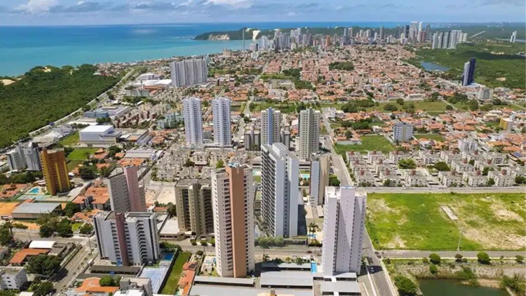Vista aérea de Natal, RN, com prédios altos, bairros residenciais e o mar ao fundo.