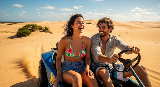 Casal sorrindo enquanto dirige um buggy azul pelas dunas douradas em dia ensolarado, aproveitando uma viagem especial de Réveillon em meio à paisagem desértica.