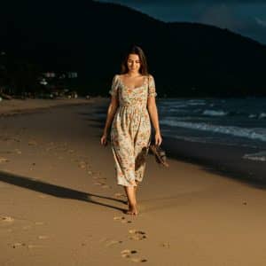 Imagem de uma mulher caminhando descalça na praia, ao entardecer, com o mar e uma montanha ao fundo.