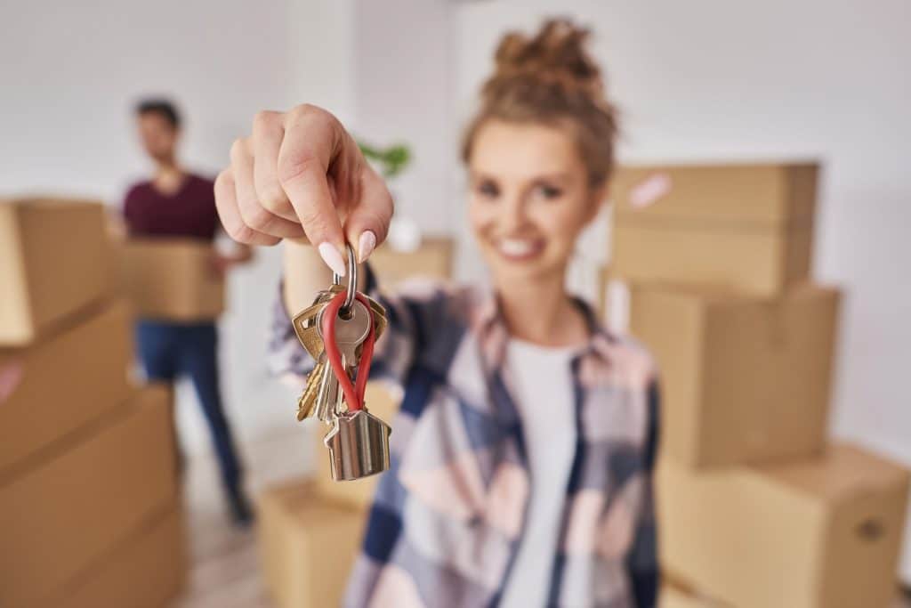 A imagem mostra uma mulher sorrindo e segurando um conjunto de chaves de apartamento, simbolizando a conquista de uma nova casa. Ao fundo, é possível ver caixas de mudança