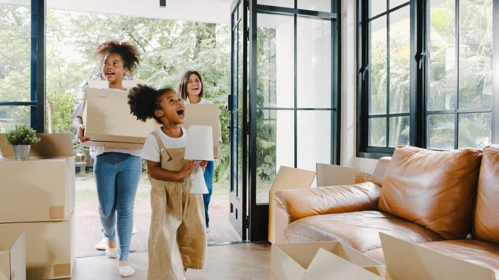 A imagem mostra uma cena alegre de mudança, com três pessoas — uma mulher e duas crianças — entrando em uma casa com caixas de papelão, simbolizando o início de uma nova etapa. A criança à frente sorri entusiasmada enquanto segura um abajur, enquanto as outras duas, mais ao fundo, também demonstram empolgação. O ambiente é iluminado por luz natural, com amplas janelas envidraçadas, piso de madeira, um sofá de couro caramelo e várias caixas espalhadas, indicando o início da organização do novo lar.