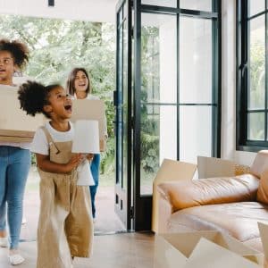 A imagem mostra uma cena alegre de mudança, com três pessoas — uma mulher e duas crianças — entrando em uma casa com caixas de papelão, simbolizando o início de uma nova etapa. A criança à frente sorri entusiasmada enquanto segura um abajur, enquanto as outras duas, mais ao fundo, também demonstram empolgação. O ambiente é iluminado por luz natural, com amplas janelas envidraçadas, piso de madeira, um sofá de couro caramelo e várias caixas espalhadas, indicando o início da organização do novo lar.