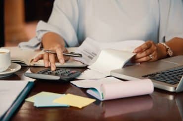 A imagem mostra uma pessoa realizando tarefas administrativas ou financeiras, utilizando simultaneamente uma calculadora, documentos impressos e um notebook. A mesa está repleta de papéis, blocos de anotações coloridos e uma xícara de café, sugerindo um ambiente de trabalho intenso e concentrado, possivelmente relacionado à contabilidade ou organização de despesas.