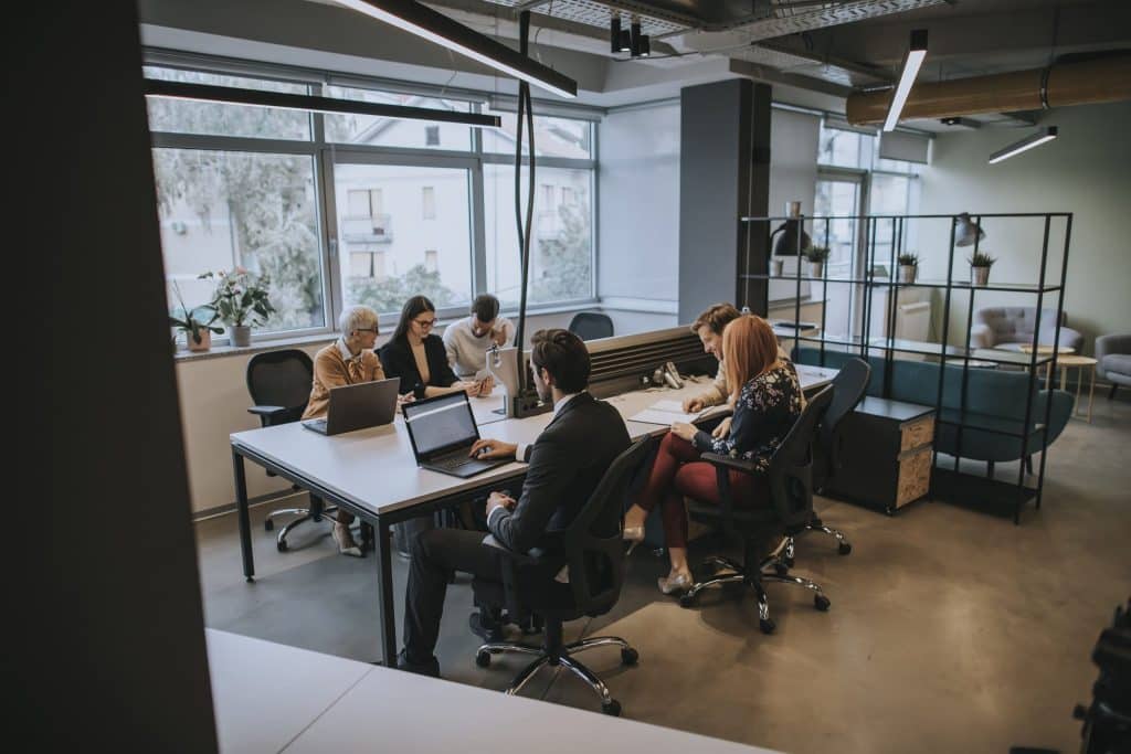 A imagem mostra um ambiente moderno de coworking, onde um grupo de pessoas está reunido em torno de mesas compartilhadas. São sete profissionais, cada um concentrado em suas tarefas, utilizando notebooks, cadernos e materiais de escritório.