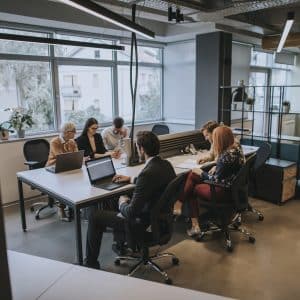 A imagem mostra um ambiente moderno de coworking, onde um grupo de pessoas está reunido em torno de mesas compartilhadas. São sete profissionais, cada um concentrado em suas tarefas, utilizando notebooks, cadernos e materiais de escritório.