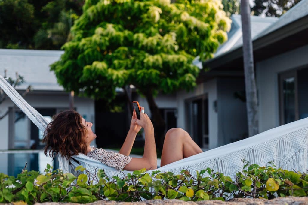 A imagem mostra uma mulher relaxando em uma rede branca em um ambiente externo cercado por vegetação, possivelmente em um imóvel de veraneio. Ela está sorrindo e usando o celular, aproveitando um momento de lazer em um local tranquilo e ensolarado, com uma casa moderna ao fundo e clima de descanso.