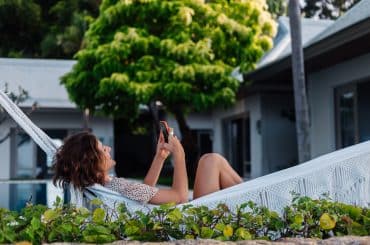 A imagem mostra uma mulher relaxando em uma rede branca em um ambiente externo cercado por vegetação, possivelmente em um imóvel de veraneio. Ela está sorrindo e usando o celular, aproveitando um momento de lazer em um local tranquilo e ensolarado, com uma casa moderna ao fundo e clima de descanso.
