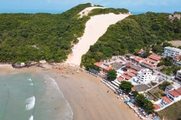 A imagem mostra uma vista aérea da Praia de Ponta Negra, em Natal (RN), destacando o famoso Morro do Careca, uma grande duna cercada por vegetação nativa. À direita, observa-se uma faixa de construções residenciais e comerciais próximas à orla, com barracas e guarda-sóis organizados na areia. O mar calmo e as pessoas aproveitando a praia completam o cenário paradisíaco e turístico do local.