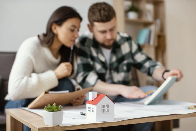 Casal analisando documentos e plantas sobre uma mesa com uma maquete de casa em destaque, representando planejamento ou compra de imóvel.