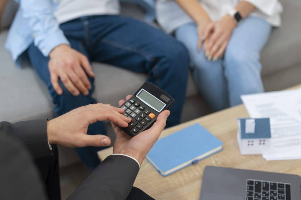 Close-up de uma pessoa segurando uma calculadora, com um casal ao fundo. Há documentos, um notebook e um bloco de notas sobre a mesa.