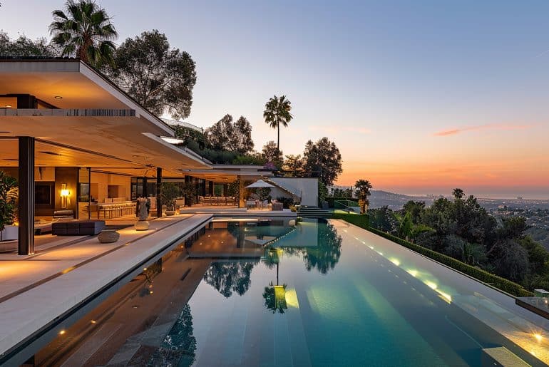 Mansão moderna com fachada aberta, piscina de borda infinita e vista panorâmica para a cidade ao entardecer, rodeada por vegetação e iluminação aconchegante.