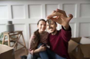 Imagem de um casal sorrindo enquanto um deles segura uma chave, em um ambiente com caixas de mudança ao fundo. O foco está na chave, com o casal ligeiramente desfocado.