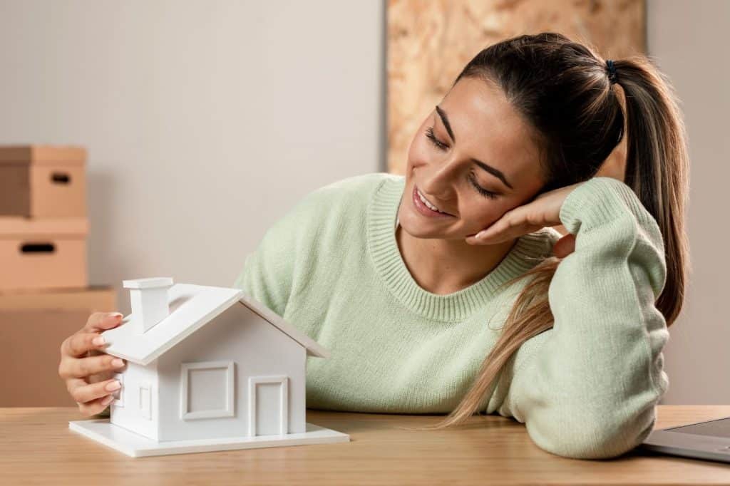 Imagem de uma mulher sorrindo e olhando com carinho para uma maquete de uma casa em um ambiente com caixas de mudança ao fundo. Ela está sentada à mesa, vestindo um suéter verde.