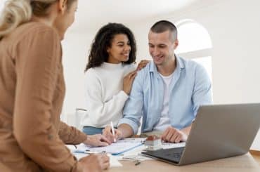 Imagem de um casal sorrindo, enquanto conversam com uma profissional em uma mesa de trabalho. A mulher está apoiando as mãos nos ombros do homem, que está sorrindo para ela. À frente deles, há documentos e um modelo de uma casa sobre a mesa, enquanto a profissional está fazendo anotações. O ambiente é claro e bem iluminado, sugerindo um ambiente confortável e profissional. A cena transmite uma sensação de cooperação e planejamento.