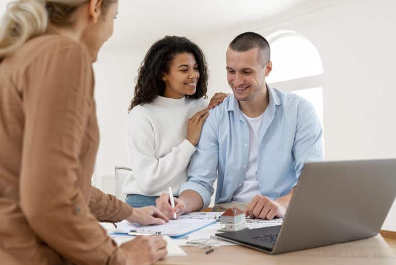 Imagem de um casal sorrindo, enquanto conversam com uma profissional em uma mesa de trabalho. A mulher está apoiando as mãos nos ombros do homem, que está sorrindo para ela. À frente deles, há documentos e um modelo de uma casa sobre a mesa, enquanto a profissional está fazendo anotações. O ambiente é claro e bem iluminado, sugerindo um ambiente confortável e profissional. A cena transmite uma sensação de cooperação e planejamento.