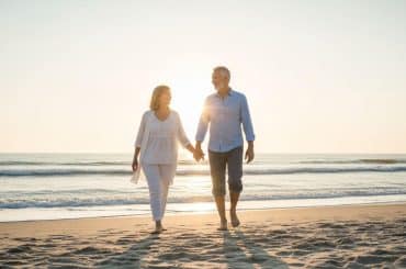 Casal de idosos caminhando de mãos dadas pela praia ao amanhecer, com o sol ao fundo iluminando a cena. Ambos estão sorrindo e vestidos com roupas leves e confortáveis.
