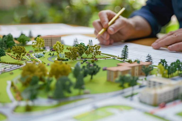 Close-up de um modelo arquitetônico de um projeto urbano, com áreas verdes, árvores e ruas, enquanto um arquiteto desenha em planos ao lado.