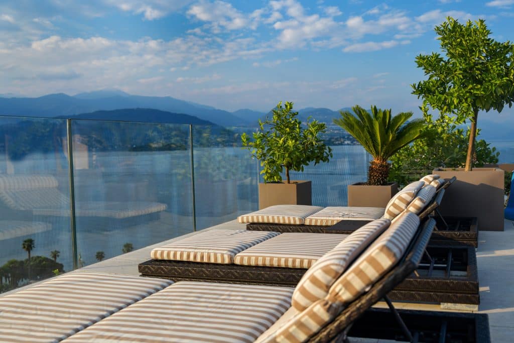 Imagem de um terraço elegante com espreguiçadeiras de listras em um ambiente ao ar livre. Ao fundo, há uma vista deslumbrante das montanhas e da água. Plantas decoram o espaço, incluindo um palmeira e árvores em grandes vasos. O vidro que cerca o local oferece uma vista panorâmica, criando um ambiente tranquilo e sofisticado. O cenário transmite um clima relaxante e convidativo.