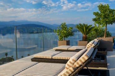 Imagem de um terraço elegante com espreguiçadeiras de listras em um ambiente ao ar livre. Ao fundo, há uma vista deslumbrante das montanhas e da água. Plantas decoram o espaço, incluindo um palmeira e árvores em grandes vasos. O vidro que cerca o local oferece uma vista panorâmica, criando um ambiente tranquilo e sofisticado. O cenário transmite um clima relaxante e convidativo.