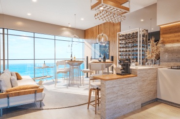 Interior de um apartamento de luxo com vista para o mar, apresentando uma área de estar com sofá e mesa de bar, além de uma cozinha moderna com adega de vinhos. A iluminação suave e os detalhes em madeira criam um ambiente aconchegante.