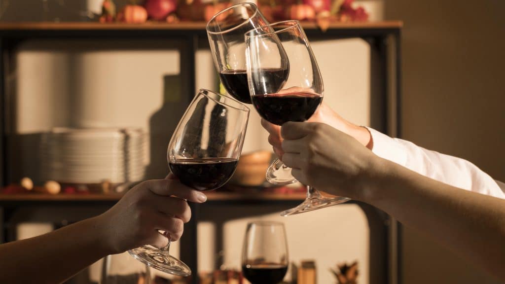 Três pessoas fazendo um brinde com taças de vinho tinto, com um ambiente acolhedor ao fundo, destacando pratos e decoração simples. A luz suave cria um clima íntimo e festivo.