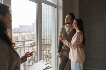 Casal abraçado observa a vista da sacada de um apartamento com corretora mostrando o imóvel.