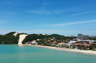 Praia com mar azul, faixa de areia e Morro do Careca ao fundo, em Natal, RN.