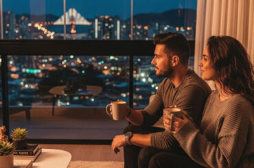 Casal sentado no sofá com canecas nas mãos, apreciando a vista noturna da cidade pela janela.