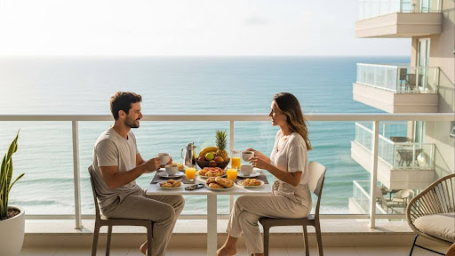 Imóveis com Vista para o Mar: O Prazer da Sofisticação Casal tomando café da manhã em varanda com vista para o mar e prédios ao fundo.