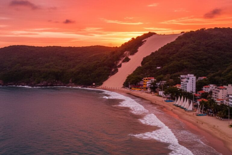 Vista aérea da Praia de Ponta Negra ao pôr do sol, com o Morro do Careca e prédios à beira-mar.