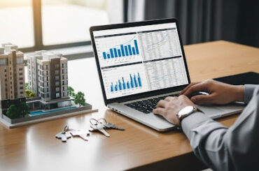 Pessoa analisando gráficos em notebook ao lado de maquete de prédio e chaves sobre a mesa.